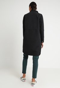 Zwarte lange jas met een kraag met knopen en een rechte silhouet. Voorzien van een achternaad, gedragen over groene broek en geruite instapschoenen.