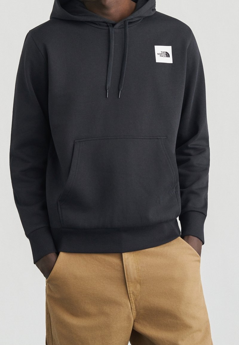 Homme portant un sweat à capuche noir The North Face avec poche avant et pantalon beige, mains partiellement dans les poches, sur un fond clair uni.