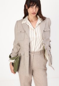 Beige fiskebein-dress med en tettsittende blazer, to brystlommer og gullknapper, kombinert med en hvit og beige stripete skjorte og grønn veske.