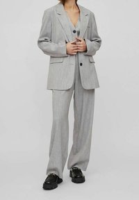 Ljusgrå kostym i en texturerad tyg, med en lös passform på kavajen som har två knappar och vida byxor. Matchad med svarta kraftiga skor.