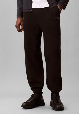 Άνδρας που φοράει μαύρο joggers Calvin Klein, μαύρα αθλητικά παπούτσια και μαύρο μπουφάν, με το ένα χέρι στην τσέπη, πάνω σε απλό γκρίζο φόντο.
