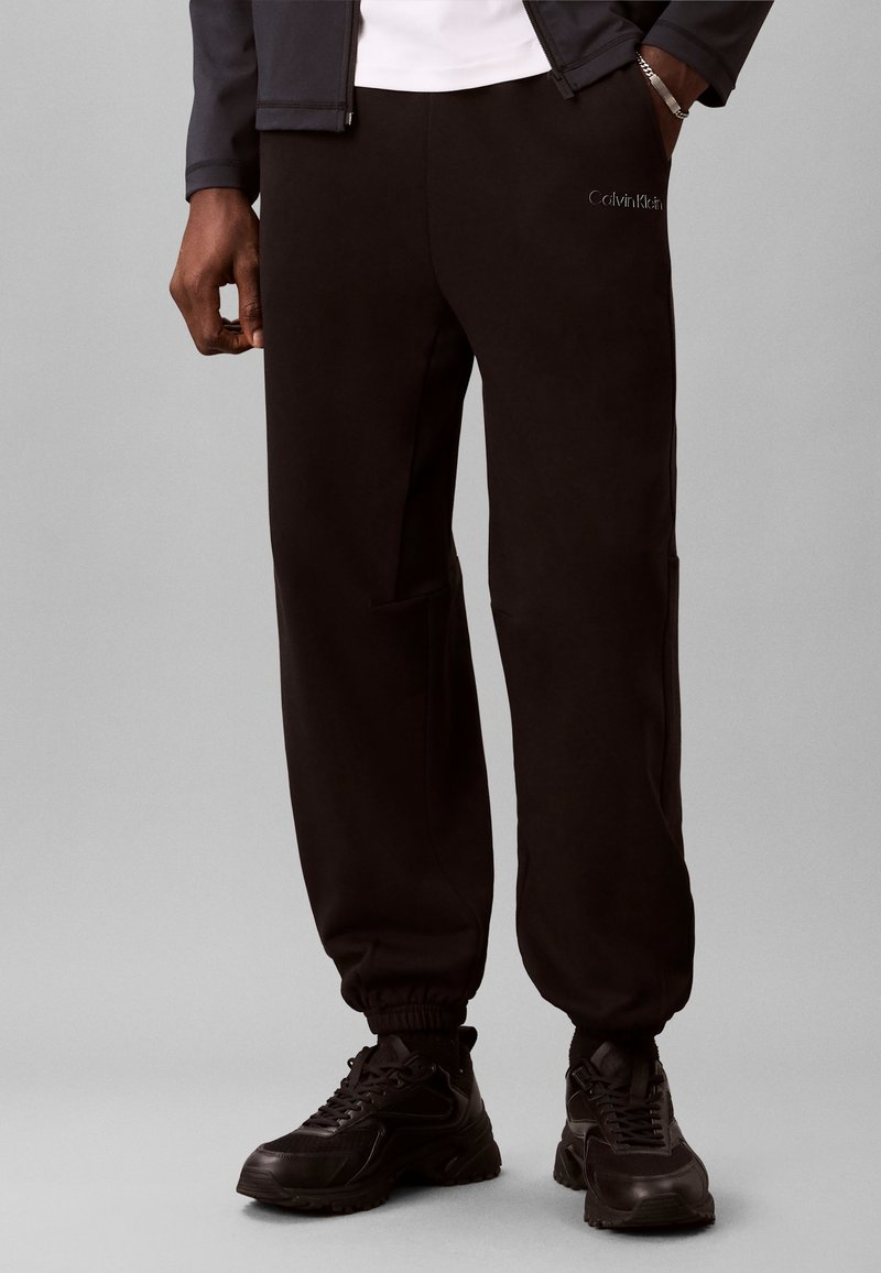 Άνδρας που φοράει μαύρο joggers Calvin Klein, μαύρα αθλητικά παπούτσια και μαύρο μπουφάν, με το ένα χέρι στην τσέπη, πάνω σε απλό γκρίζο φόντο.