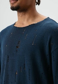 Marineblauwe, gestructureerde trui met een losse pasvorm en grote, onregelmatig verdeelde gaten. Versleten halslijn en mouwen dragen bij aan het verweerde ontwerp.