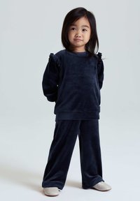 Marineblauw velours trainingspak met lange mouwen met ruches en losse broekspijpen. Gladde textuur, eenvoudig ontwerp en neutraal schoeisel.