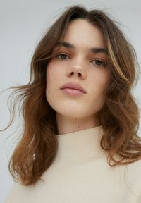Close-up van een persoon met middellang golvend bruin haar, die een lichtbeige hoge hals trui draagt. De huid vertoont een natuurlijke textuur met sproeten.