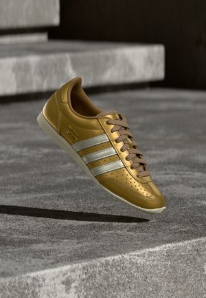 Złoty skórzany sneaker z trzema białymi paskami, sznurowanym zapięciem, perforowanymi detalami i jasnokolorową gumową podeszwą.