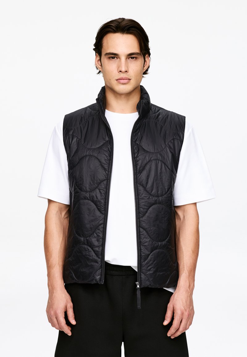 Jeune homme portant un gilet matelassé noir à fermeture éclair sur une chemise blanche à manches courtes et un pantalon noir, debout devant un fond uni.