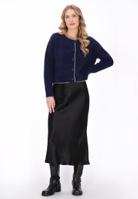 Kobieta w granatowym, puszystym kardiganie zapinanym na guziki, czarnej satynowej spódnicy midi i czarnych skórzanych kozaczkach, stojąca z jedną ręką na biodrze.