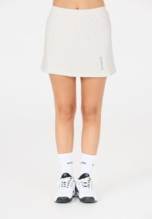 Ljus beige sportkjol med en elastisk midja och tryckt logotyp. Kombineras med vita strumpor och vit-svarta sportskor. Slät textur.