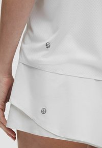 Valge sportlik särk, millel on tekstuuriga viimistlus, kihiline disain ja ümar alläär. Väike hall logo on nähtav alumise serva lähedal.