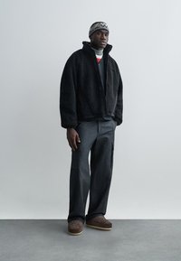 Veste polaire noire à col montant, associée à un pantalon cargo foncé et des chaussures marron à enfiler. Bonnet noir et blanc à motifs sur la tête.