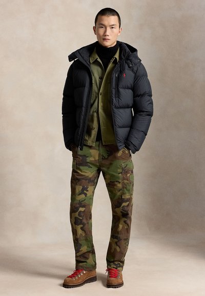 Young man standing wearing a black puffer jacket, green shirt, camouflage cargo pants, and brown hiking boots with red laces.