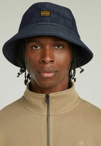 Cappello a secchiello in denim blu navy con una larga tesa, caratterizzato da un'etichetta gialla G-Star sul davanti. Indossato con una felpa beige con zip e colletto.