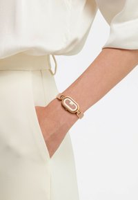 Armband aus Roségold mit einem rechteckigen Gehäuse, das einen strukturierten Mesh-Hintergrund aufweist. Weicher beiger Riemen mit runden Hardware-Akzenten.