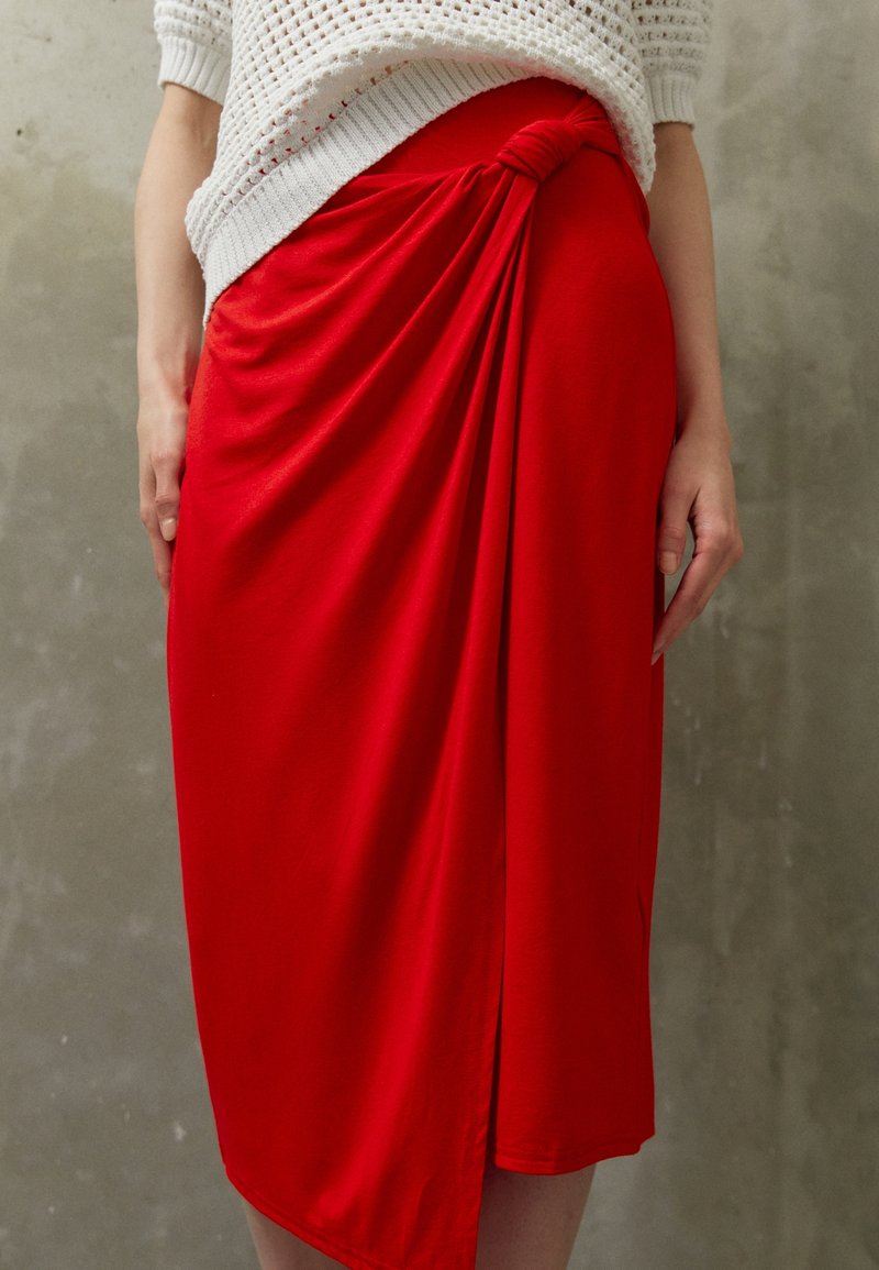 Red knee-length skirt made of soft fabric, featuring a draped design with a knotted detail at the waist and a side slit for ease of movement.