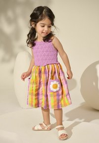 Robe à corsage tricoté violet avec une jupe à carreaux multicolores, dotée d'une poche brodée de fleurs. Des sandales blanches complètent le look.