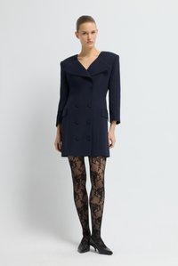 Cappotto doppiopetto blu con ampi rever e tasche laterali, abbinato a collant neri a fantasia floreale e tacchi neri.