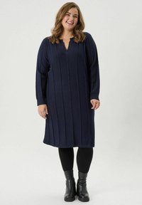 Tumesinist kudumist kleit V-dekolteega ja tekstuursete horisontaalsete triipudega. Pikad varrukad, põlvepikkune lõige, koos mustade sukkpükste ja pahkluu saapadega.