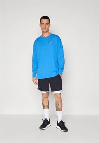 Camisa azul de manga comprida com logótipo, combinada com calções pretos e meias brancas. Apresenta tatuagens visíveis nas pernas e sapatos atléticos pretos.