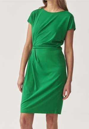 Femme portant une robe verte longueur genou avec des manches courtes et un détail froncé à la taille, debout devant un fond uni.