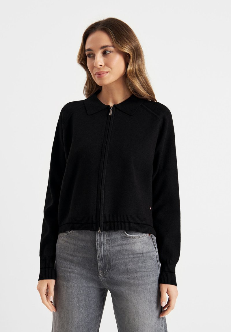 Zwarte zip-up sweater met een kraag, geribbelde onderrand en manchetten, gemaakt van een zachte stof. Draagt met grijze jeans, voorzien van een subtiele logodetail.