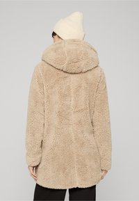 Persoon die een beige, donzige jas met capuchon en een crèmekleurige gebreide muts draagt, van achteren gezien tegen een effen lichtgrijze achtergrond.