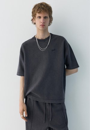 Giovane uomo con capelli castano chiaro che indossa una maglietta scura oversize, pantaloni abbinati, collana con catenina d'argento e anello, in piedi contro una parete semplice.