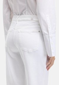 Białe dżinsy z denimu mają wysoki stan, tylne kieszenie oraz delikatne detale szwów. Częściowo widoczna jest dopasowana biała koszula.