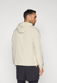 Világos bézs kapucnis dzseki húzózsinóros alsó résszel és elasztikus mandzsettákkal, sima, vízlepergető anyagból készült. Hátsó nézet.