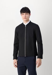 Must musta zip-up jakk, millel on ümar kaelus, valmistatud siledast kangast. Omab küljetaskuid ja on sobiva lõikega, kombineeritud valge särkiga all.