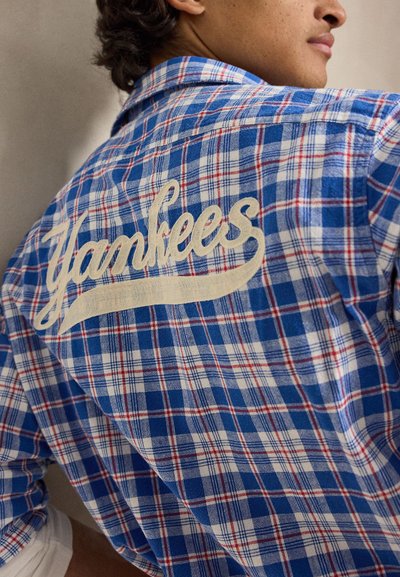 Osoba u plavoj, bijeloj i crvenoj kockastoj košulji s izvezenim natpisom „Yankees“ u kremastom pismu na leđima, lagano okrenuta od kamere.
