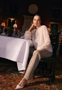 Woman in textured white blouse and plaid trousers sits at a table with a white tablecloth, surrounded by glassware and candles.