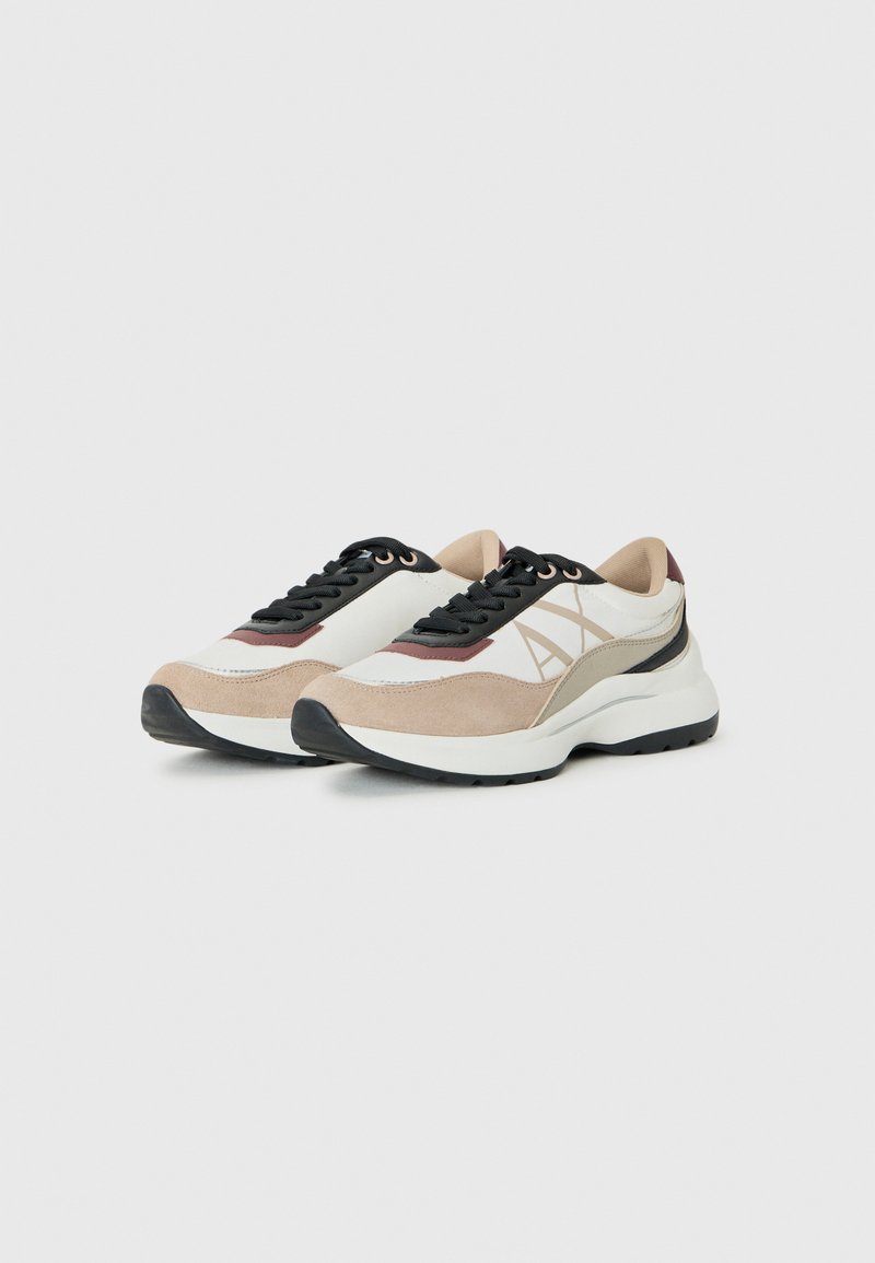 Zapatillas deportivas ligeras en beige, blanco y ante marrón con suela negra. Presentan un diseño con cordones y un detalle de logo en el lateral.