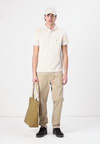 Creme polo shirt, beige bukser, sorte sneakers og bærer en lysbrun tote bag. Mand står mod en ensfarvet hvid baggrund.