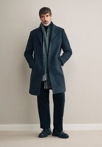 Cappotto in lana grigio scuro con rever a incavo, design monopetto e tasche laterali, indossato sopra una sciarpa verde. Pantaloni e scarpe nere.