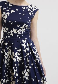 Navy dress with white butterfly print, featuring a fitted bodice and pleated skirt. Made from fabric with a smooth texture and cap sleeves.