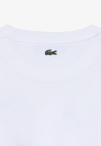 Balta medvilnės marškinėliai su apvaliu iškirpimu, kuriuose ant nugaros, netoli kaklo, išsiuvinėtas mažas žalias krokodilo logotipas.