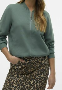 Vihreä vetoketjullinen collegepaita, jossa on resorit hihansuissa ja kauluksessa; yhdistetty leopardikuvioiseen hameeseen, jossa on taskut ja hieman A-linjainen malli.