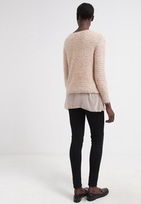 Ljusrosa fluffig tröja med en texturerad stickning, lagd över en genomskinlig, ljus taupe-blus, tillsammans med svarta slim-fit byxor och svarta platta skor.