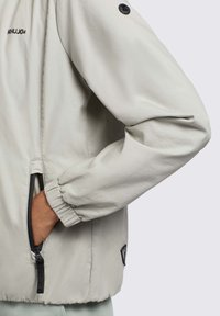 Lichtgrijze jas met een ritssluiting aan de voorkant, elastische boorden, een minimalistisch ontwerp en een logo detail op de schouder. Glad stoffen textuur.