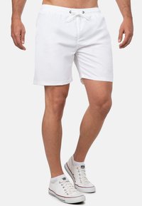 Pantaloni da nuoto bianchi con cintura elastica, cordoncino e tasche laterali. Tessuto liscio con un design leggermente aderente, taglio sopra il ginocchio.