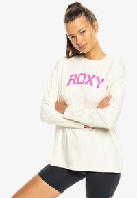 Joven mujer con el cabello recogido, vistiendo una camisa blanca de manga larga con "ROXY" en rosa y negro, y pantalones cortos de ciclismo, con los brazos cruzados.