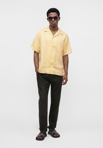 Homme portant des lunettes de soleil foncées, une chemise jaune à manches courtes avec boutons, un pantalon noir et des sandales noires, debout devant un fond clair uni.