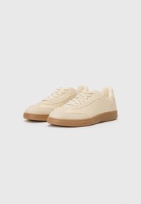 Beige sneakers with a textured upper and tonal stitching, featuring a rounded toe and rubber gum sole. Lace-up design for secure fit.