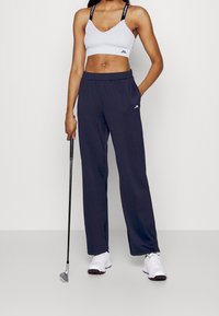 Bílá žebrovaná sportovní podprsenka a tmavě modré široké kalhoty z lehké látky s elastickým pasem a logem. Drží golfovou hůl.