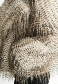 Close-up of a textured beige and brown spotted fur coat with long, soft fibers worn over black pants.