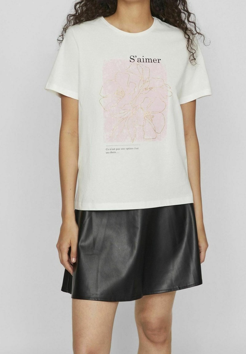 Camiseta de algodón blanca con un gráfico floral rosa con contorno dorado y texto. Combinada con una falda de cuero negra.