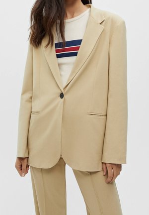 Woman wearing a beige suit with a single-button blazer over a white shirt featuring navy blue and red horizontal stripes.