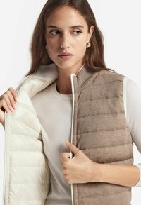 Bruine gewatteerde vest met een beige binnenkant, geribde kraag en witte rits. Draagt over een lichte trui, waarbij de textuur en gelaagde ontwerp worden getoond.