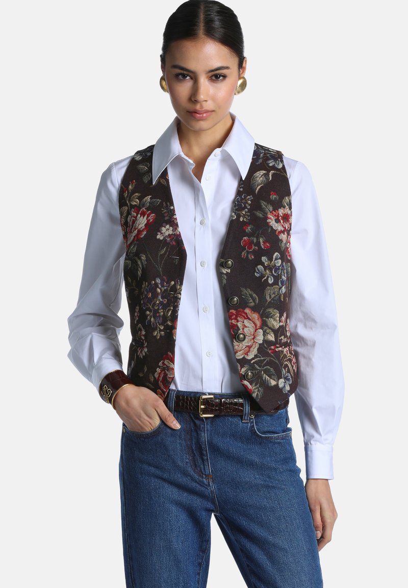 Femme portant une chemise blanche boutonnée, un gilet sombre à motif floral, un jean bleu, des boucles d'oreilles dorées et une ceinture en cuir marron avec une boucle dorée.